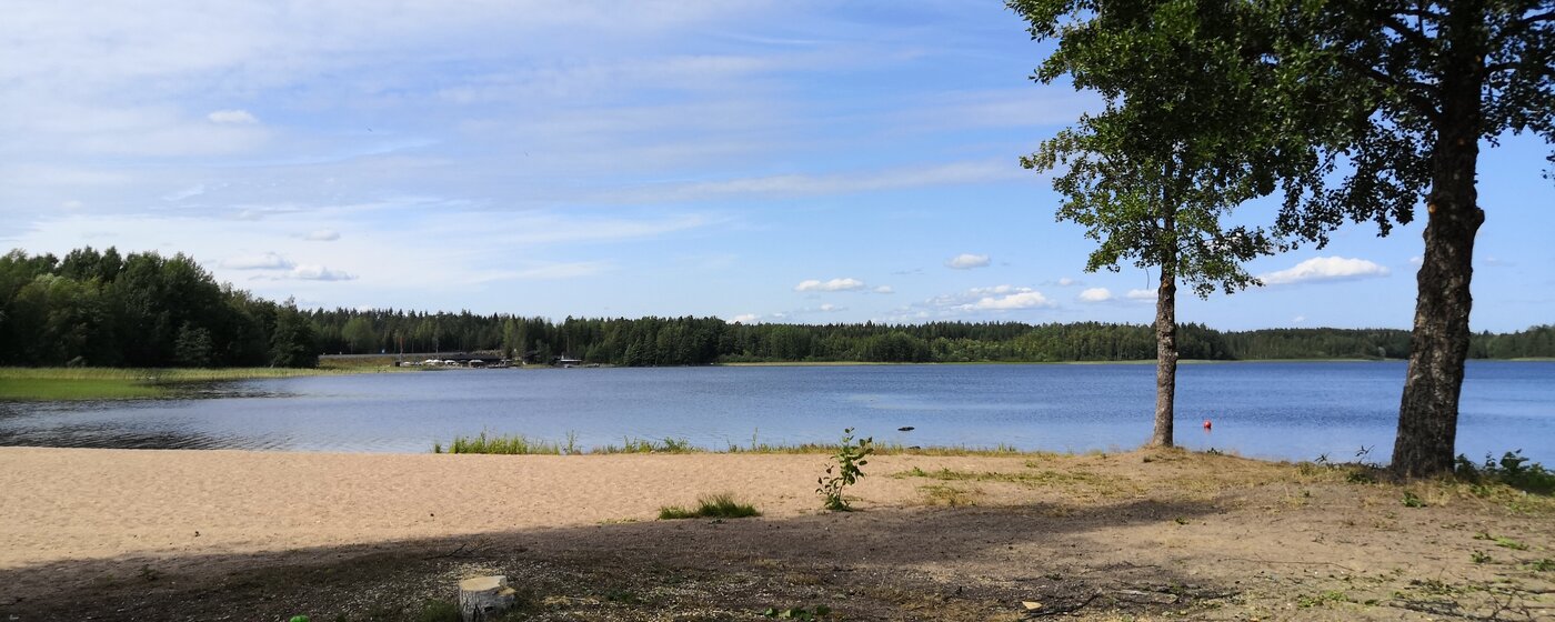 Käyrälammen hiekkaranta ja tyyni järvenselkä aurinkoisena kesäpäivänä Kouvolassa, rannan puut kehystävät näkymää.