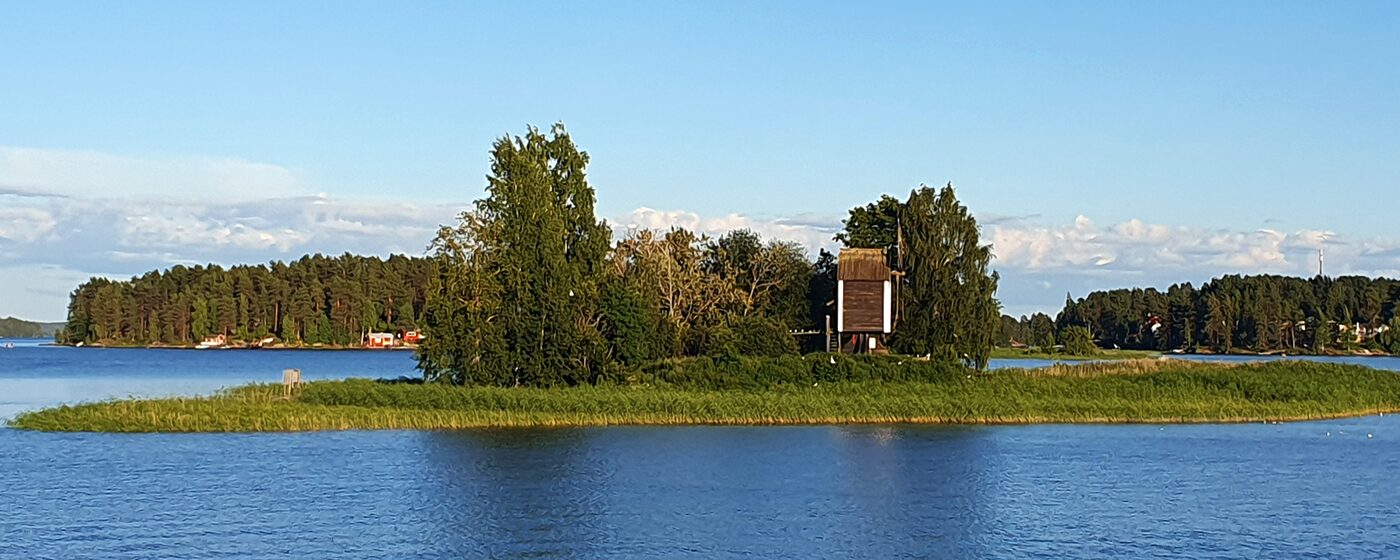 Vasikkasaaren vehreä saari ja vanha tuulimylly Kallavedellä Kuopiossa aurinkoisena kesäiltana, ympärillä sinistä järvenselkää.