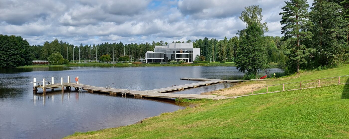 Pankalammen kesäinen ranta Mikkelissä: laiturit tyynellä vedellä, vihreä nurmiranta ja valkoinen Mikaelin konserttitalo vastarannalla.