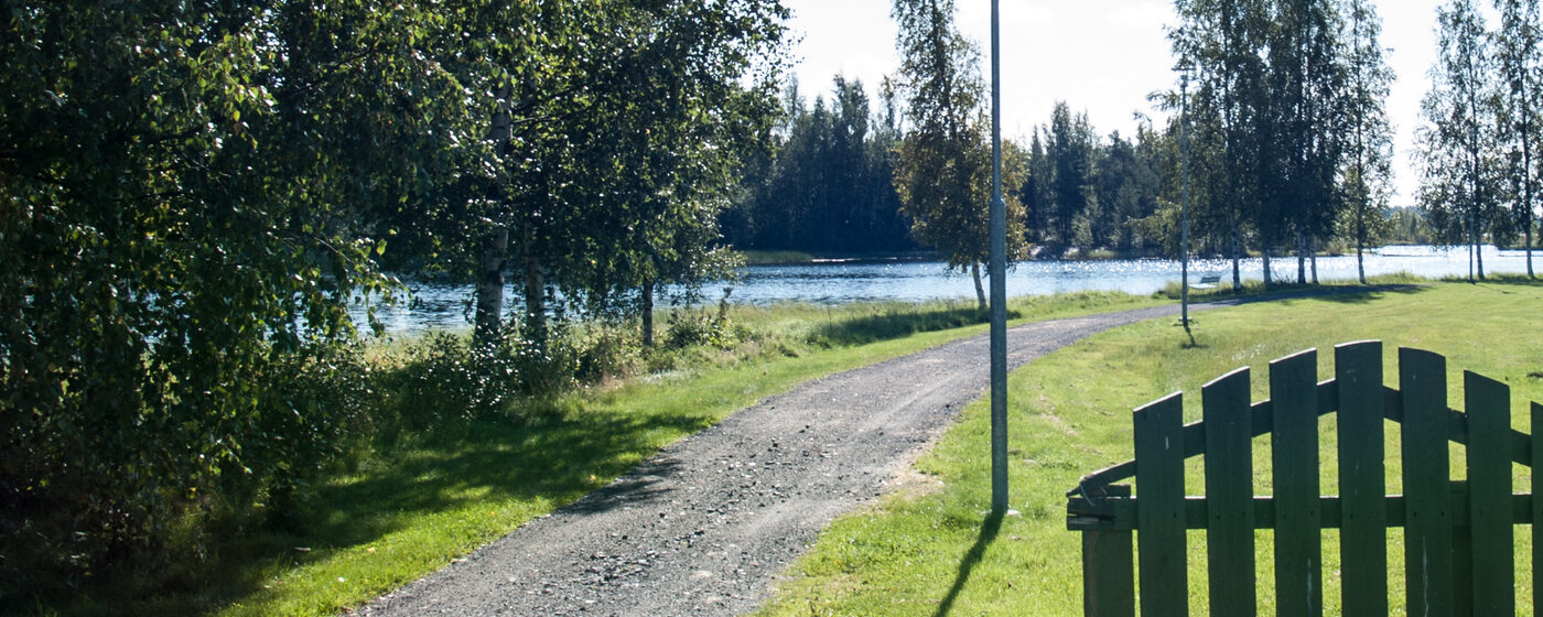 Sorapolku johtaa kesäisen puiston läpi Kyrkösjärven tekojärven rantaan Seinäjoen Huhtalassa; oikealla puinen aitaportti, vasemmalla nurmea ja koivuja.